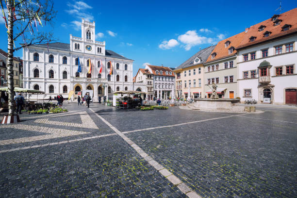 Sunny view of the bustling market square in Weimar, Germany, featuring historic buildings, flags, cobblestone streets, and visitors.