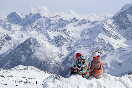 In the background are the Caucasus Mountains, Mount Ushba, and the border with Georgia. View point. A couple of tourists sit in the snow and admire the view of the mountains. Winter bright clothes. Ski resort of the Elbrus region. Kabardino-Balkaria, Russia 202
4