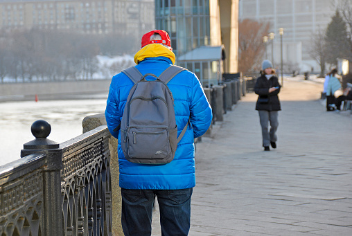 Russia, Moscow - March 18, 2024: An elderly man in casual bright clothes stands on the street with a backpack. He is wearing jeans, a blue jacket, a red baseball cap and a yellow hoodie. View of a person from the back. The street background is blurred.