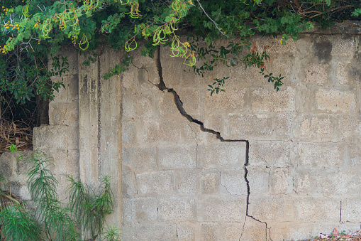 Crack in a concrete fence made of cement blocks, appeared as a result of soil shrinkage in area of pole and as a result of rupture and partial destruction. Defects in construction and their causes.