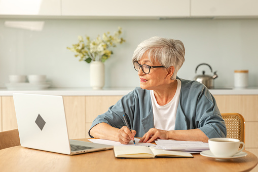 Middle aged senior woman using laptop computer writing notes at home. Focused mature old businesswoman work in home office making written records doing online work. Mature boss professional worker