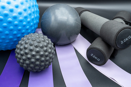 Wellness and fitness tools: fitness bands in different colors, exercise ball, massage ball, weight ball and two 2kg dumbbells. Close-up on dark background.