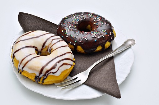 glazed donut and chocolate donut with sugar sprinkles on white plate with brown napkin and fork, close up, isolated on white background