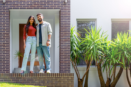 Couple standing proudly in front of their new home. They are both wearing casual clothes. They are looking at the camera and smiling. The house is contemporary with a brick facade. The front door is also visible. Copy space