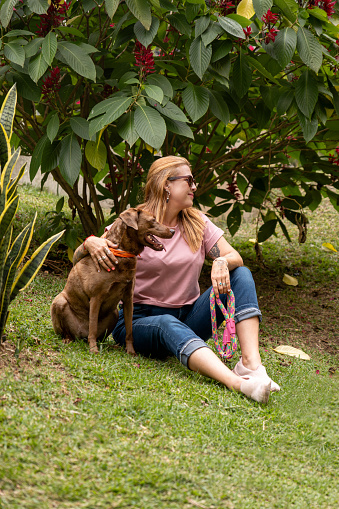 Vertical shot of a cheerful middle-aged woman with her dog, both looking to the side, seated on the grass surrounded by plants and flowers.