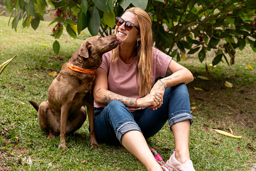 Woman in her 40s wearing sunglasses with a joyful expression receives a lick from her pet while they both sit on the grass, enjoying a sunny day in the park.