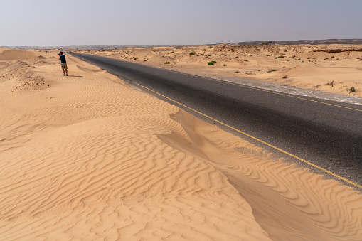 One men in distance walking sand dune close to the asphalt road, sand almost covering the road, Wahiba rippled desert in Oman.