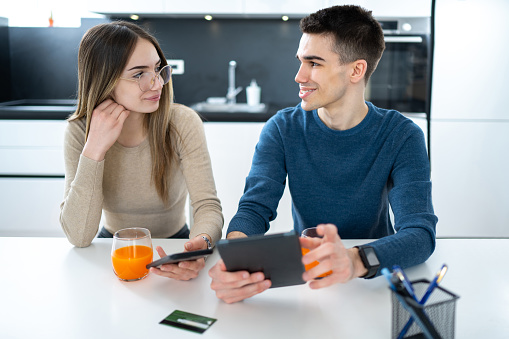 Beautiful young teenage couple with tablet and smartphone talking and looking each other at home