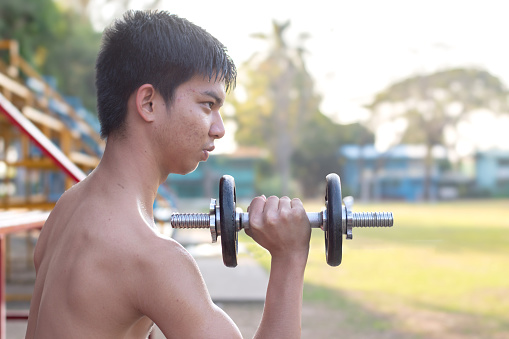 Asian cute teenboy doing exercise with dumbbells in outdoor park in late afternoon of the day.