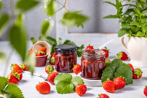 Fresh homemade strawberry jam in glass jar on a light background. Several fresh berries, green strawberries leaf and flowers are near it.