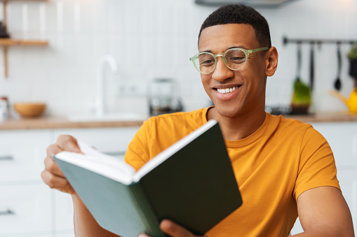 Smiling attractive African American man wearing stylish eyeglasses holding and reading book while sitting in kitchen at home. Handsome young businessman is planning project. Concept of hobby, relaxing