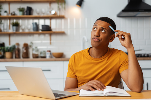 Thinking pensive African American man, freelancer working using laptop taking notes attractive businessman writing in diary, student preparing for exam remote work at home. Concept of online education