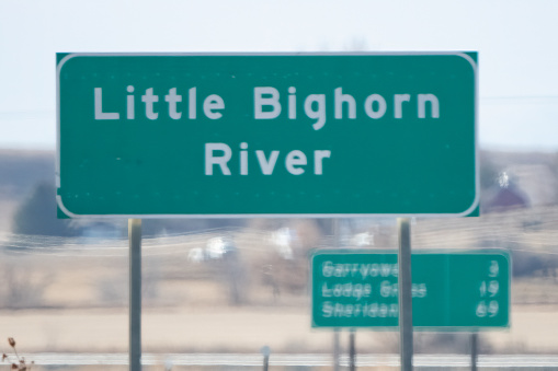 Highway sign marking the Little Bighorn River at the Little Bighorn Battlefield National Monument in winter in southeastern Montana, in western USA of North America. Commemorates the Battle of the Little Big Horn which took place in June 1876. The Little Big Horn river runs across the top of the photo. It is lined with trees.
