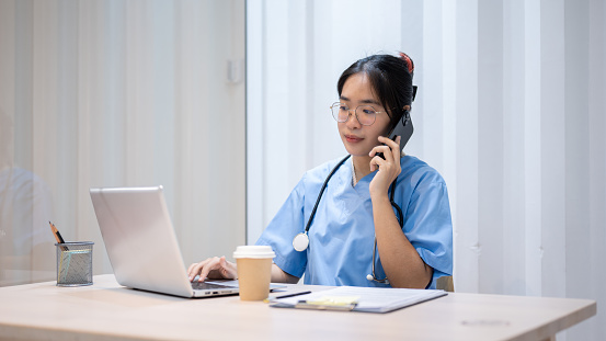 A professional and hardworking Asian female doctor is multitasking, talking on the phone with someone while reading medical cases on laptop in her office at the hospital.