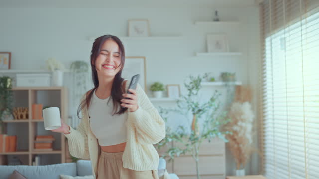 Happy carefree asian woman holding coffee cup and mobile smartphone have fun moving listening to music relax in living room, She dance enjoy leisure weekend at home.