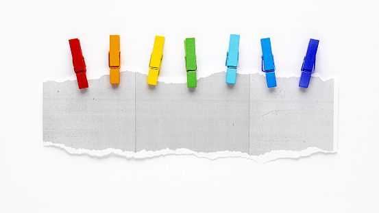 A row of vibrant clothespins attached to a torn white copier paper on white background.