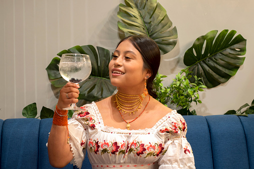 A cheerful young indigenous woman is sitting on a blue sofa. He is holding a glass of wine and appears to be participating in a social event, smiling and looking to the side