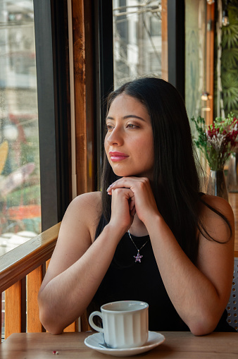 A serene young woman enjoys a moment of peaceful contemplation by the window of a cozy cafe. As she gazes thoughtfully outside, the warm ambiance of the cafe embraces her, accentuated by soft natural light. The cup of coffee before her suggests a break in her explorations, perhaps a quiet interval in a day full of discovery and adventure.
