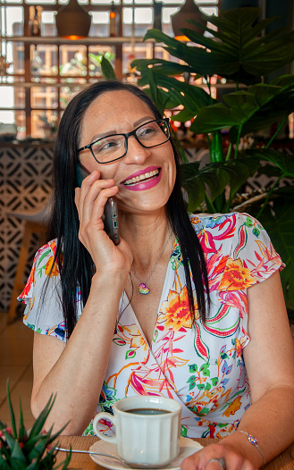 A joyful woman in a floral blouse and glasses is engaged in a pleasant conversation on her smartphone. She sits at a wooden table in a cozy cafe, with a cup of coffee, smiling warmly. Lush green plants and intricate patterns add to the relaxed ambiance.