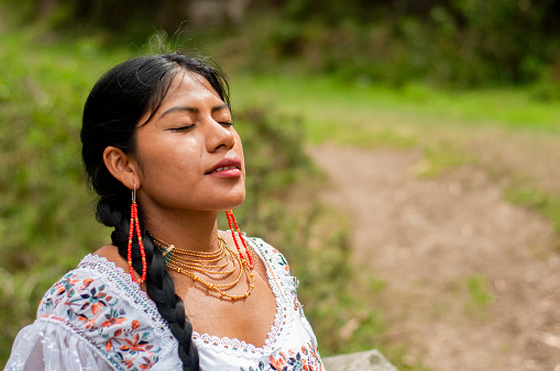 A young woman adorned in traditional embroidered clothing is captured in a tranquil moment with her eyes closed, basking in the serenity of the surrounding greenery. Her traditional earrings and necklace complement the intricate patterns on her dress while she takes a peaceful breath in the natural park setting at dusk.