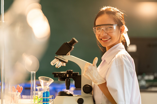 Poster image for website of a smiling Asian female chemist. Raise hand, like symbol, use microscope, work in company, business, medical industry, in biological lab, ministry of science