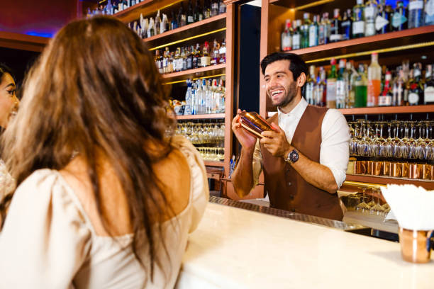 istock Caucasian professional bartender or mixologist making a cocktail for women at a bar. Attractive barman mixes liquor ingredients with a cocktail shaker with a smile. Bartender at night club restaurant. 2095708556