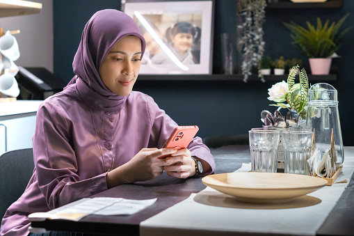 woman in hijab sitting at dining table playing with cellphone, showing absent-minded or smiling expression, as if waiting for someone