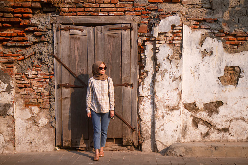 happy asian muslim woman wearing sunglasses standing in front of wooden door with old grunge brick wall while roaming around kota lama or old city area, Semarang, Central Java , Indonesia