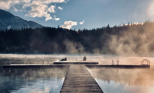 View down dock over lake to mountains, forest and lofty clouds in morning light and mist. Couple at end of dock