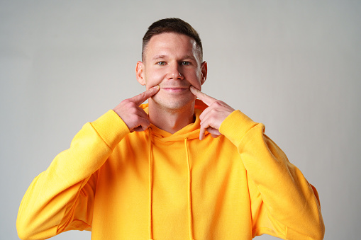 Young man in yellow hoodie smiling and pointing to cheeks on gray background in studio