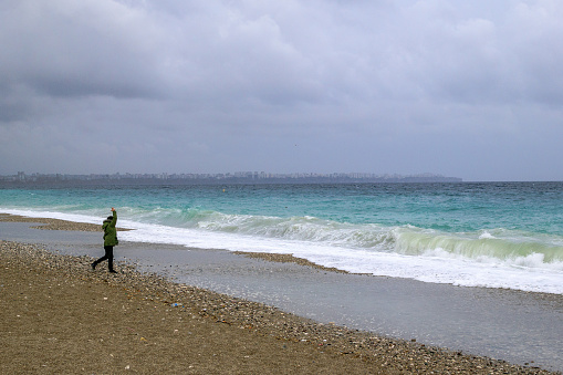 Antalya, Turkey - March 05, 2023: Unrecognizable man is walking near to stormy sea with big waves and city on background.