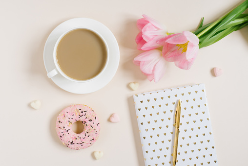 Trendy stylish concept for a female blogger, freelancer: pink diary, notebook, Donat, pink tulips and an cup of coffee  on beige background. Working from home place