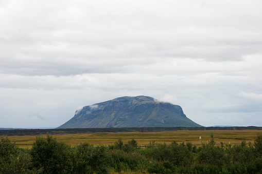 On a cloudy day in Iceland, a massive mountain looms majestically over vast agricultural plains, stretching across the horizon in a breathtaking display of natural grandeur, as the ever-changing sky casts shifting shadows and moods upon the rugged landscape below.