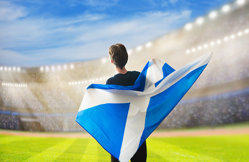 Scotland football supporter on stadium. Scottish fans on soccer pitch watching team play. Group of Scotland supporters with flag and national jersey cheering for Scotland. Championship game. Go Scotland!