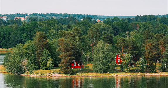 Panoramic View On Many Red Swedish Wooden Sauna Logs Cabins Houses On Island Coast In Summer Cloudy Day. Swedish Old Tradition Wooden Houses. Bold Colors. Travel to Sweden.