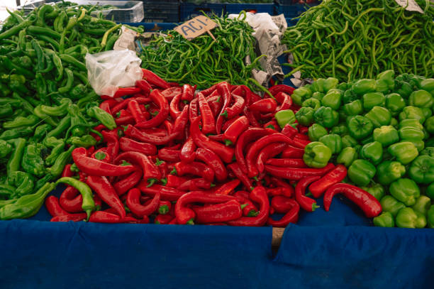 Adult woman wearing a red overcoat, sunglasses and a blue scarf shopping in farmer's market. She is choosing green peppers. She has black hair. Selective focus on model. Shot in daylight with a full frame DSLR camera.