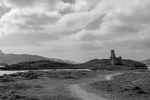 A black and white image capturing the serene landscape of Ardvreck Castle ruins, nestled amidst the rugged terrain of Scotland. The winding path leads the eye towards the historic structure