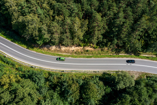 Top down view of two cars driving down a winding road through a forest in summer.