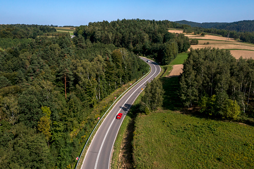 Red car driving down a winding road among forests and fields seen from above.
