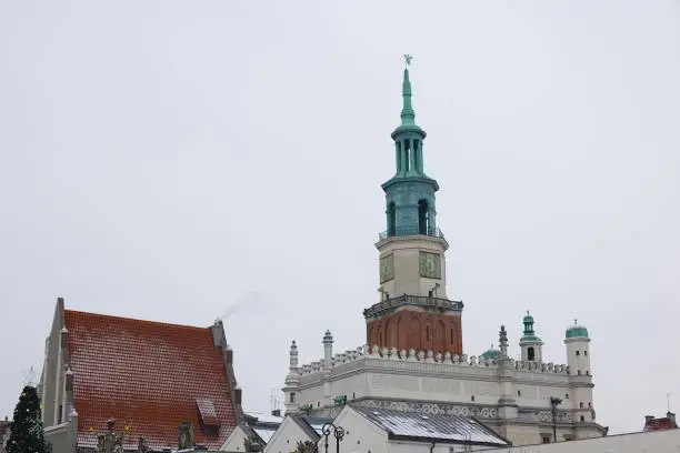 A photo of the old market square in Pozna, Poland, taken in January during winter. Snow-covered rooftops with the historic town hall against a white sky backdrop.