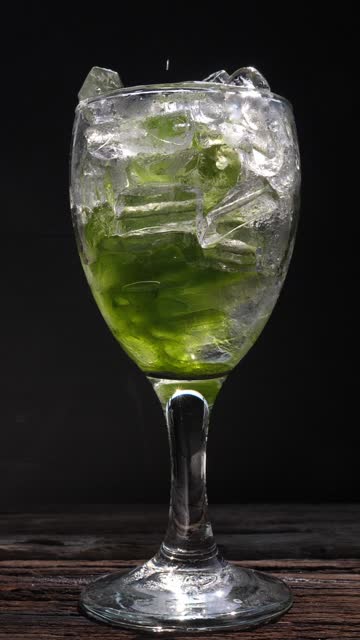 Pouring kiwi syrup into the wineglass with ice cubes against a black background, close-up and slow-motion