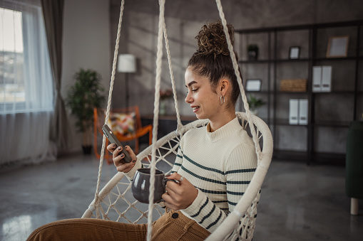 A young beautiful woman is sitting in a cozy wicker swing in her living room and enjoying her morning coffee
