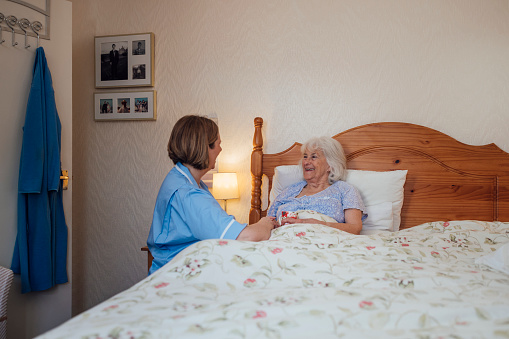 Senior woman lying in bed being helped by a home career in the North East of England. The nurse has given the woman a cup of tea, starting her morning routine.