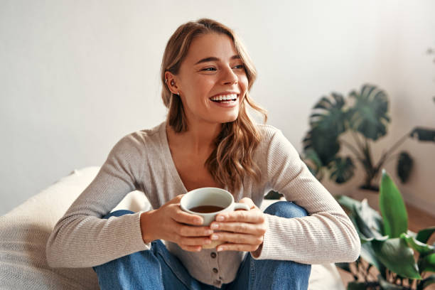 Woman at home Young woman with a cup of hot coffee or tea sitting on a sofa in a cozy living room at home relaxing and unwinding on a weekend day. woman lifestyle stock pictures, royalty-free photos & images