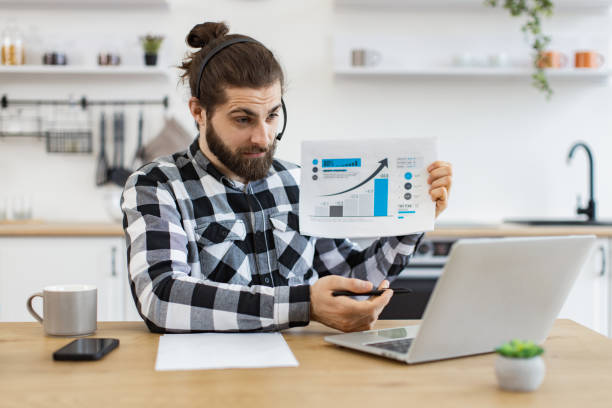 Qualified data analyst demonstrating information about business growth. Qualified data analyst demonstrating information about business growth in kitchen interior. Attractive male holding paper with charts while speaking during online meeting via computer at home. qualified report stock pictures, royalty-free photos & images