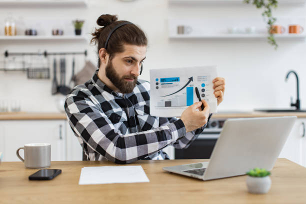 Qualified data analyst demonstrating information about business growth. Qualified data analyst demonstrating information about business growth in kitchen interior. Attractive male holding paper with charts while speaking during online meeting via computer at home. qualified report stock pictures, royalty-free photos & images