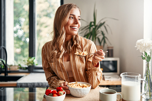 Young happy woman sitting at the table in the kitchen, eating cereal with milk, strawberries and drinking coffee. A woman is having breakfast in a cozy kitchen.