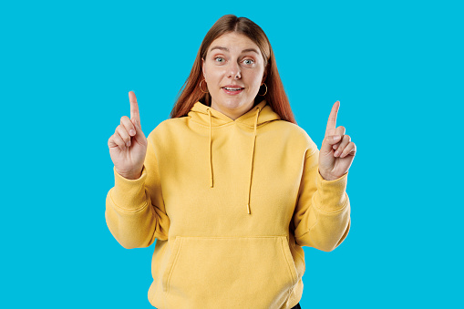 Cheerful female model pointing fingers up to shop promotion banner, smiling camera, studio portrait. 30s Woman pointing fingers at copy space isolated over blue background