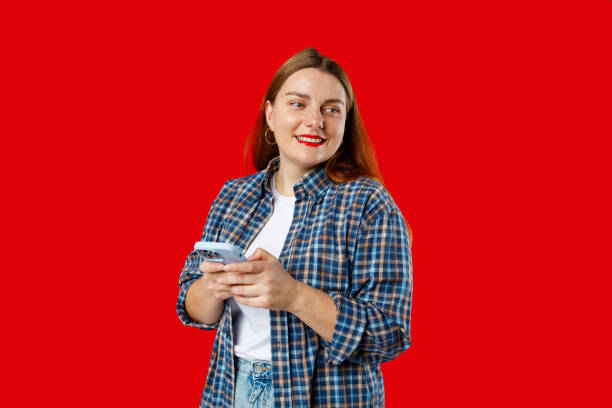 istock Smiling young brunette woman with smart phone posing isolated on red background, studio portrait. Cute beautiful young woman chatting by mobile phone 2096170353