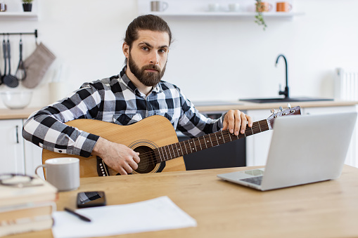 Portrait of handsome bearded adult posing with six-string guitar while enjoying pleasant pastime at home. Smiling Caucasian man learning to play instrument using online lessons on laptop.